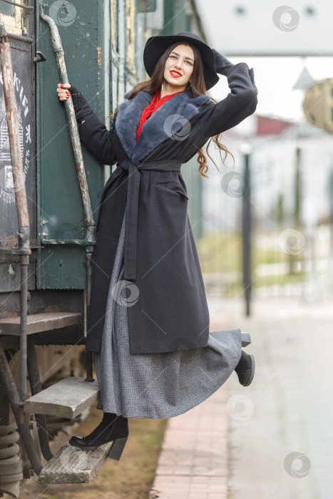Скачать Красивая девушка в черном пальто и шляпе возле старого паровоза и больших железных колес. Молодая женщина с длинными темными волосами. Винтажный портрет прошлого века, ретро-путешествие. фотосток Ozero