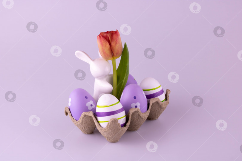 Скачать Белый керамический кролик с тюльпаном в коробке для яиц, рядом с ним лежат пасхальные яйца фотосток Ozero