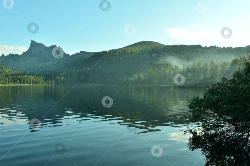 Скачать Отражение высокой скалы в большом живописном озере, окруженном густым лесом, над которым теплым летним вечером поднимается дым от костра. фотосток Ozero