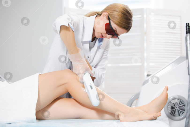 Скачать Фотография симпатичной женщины-врача в специальных защитных очках, которая проводит процедуру лазерной эпиляции, удаление волос на ногах современными методами фотосток Ozero