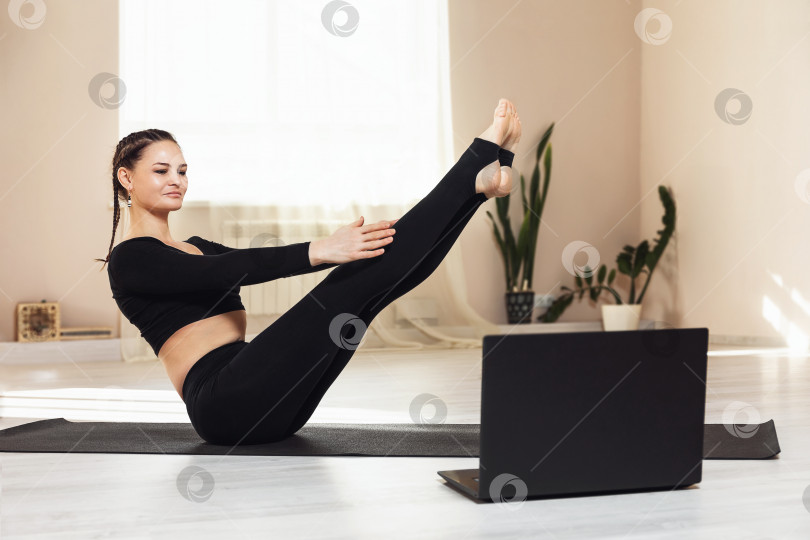 Скачать Женщина в черной спортивной одежде практикует йогу, выполняя онлайн-упражнение Наукасана, поза лодки. Концепция на тему дистанционного обучения из дома фотосток Ozero