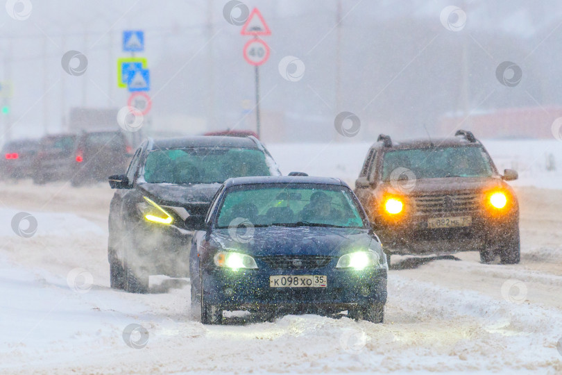 Скачать Череповец, Россия, 27 ноября 2023 года, автомобили движутся по заснеженной дороге на городской улице во время сильного снегопада зимой. Затруднение движения из-за снега и метели. Прогноз погоды, пробка на дорогах фотосток Ozero
