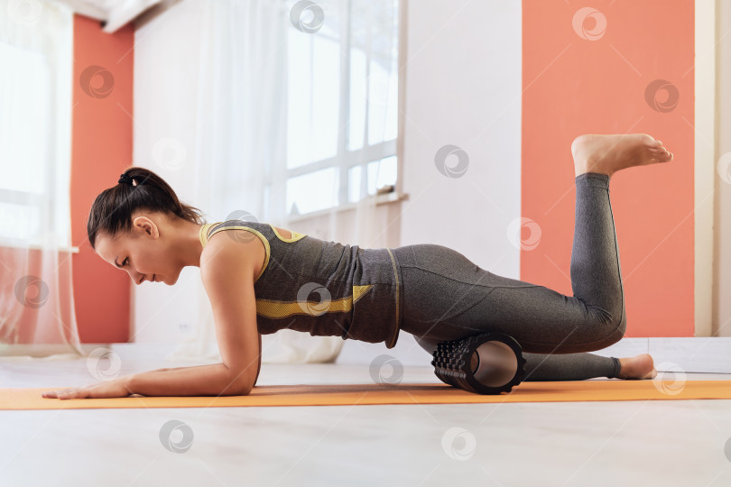 Скачать Брюнетка в спортивной одежде выполняет миофасциальный массаж бедра валиком, лежа на гимнастическом коврике. Концепция на тему восстановления мышц после травмы фотосток Ozero
