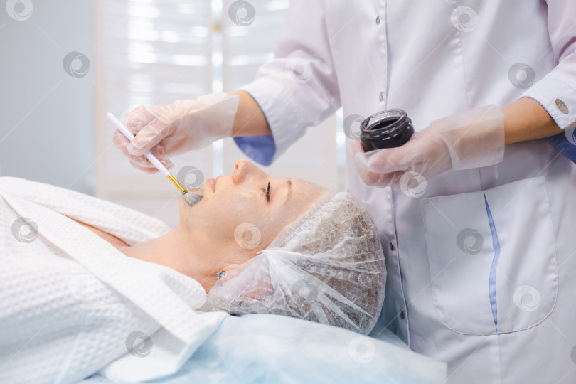 Скачать Вид сбоку на руки косметолога, наносящего углеродный наногель на лицо молодой пациентки с помощью кисти, готовящейся к процедурам углеродного пилинга фотосток Ozero