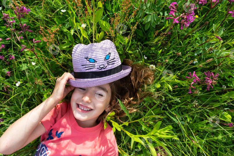 Скачать Портрет ребенка в шляпе с закрытым летом лицом, лежащего в траве и полевых цветах. Шляпа с глазами и ушами, как у кошки, летнее время, свобода фотосток Ozero