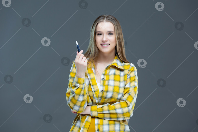 Скачать Студийный снимок привлекательной улыбающейся девушки, смотрящей в камеру и держащей электронную сигарету в поднятой руке, выделенный на серо-голубом фоне фотосток Ozero