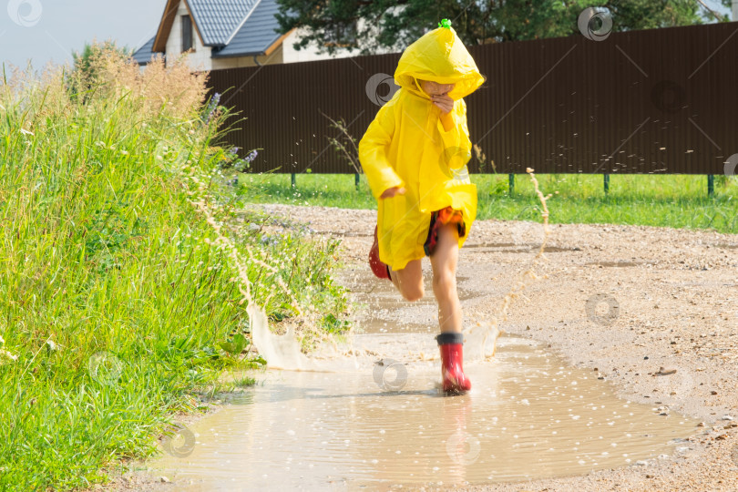 Скачать Девочка в красных резиновых сапогах и желтом дождевике бежит по лужам после дождя в деревне. Летнее время, свобода, детство фотосток Ozero