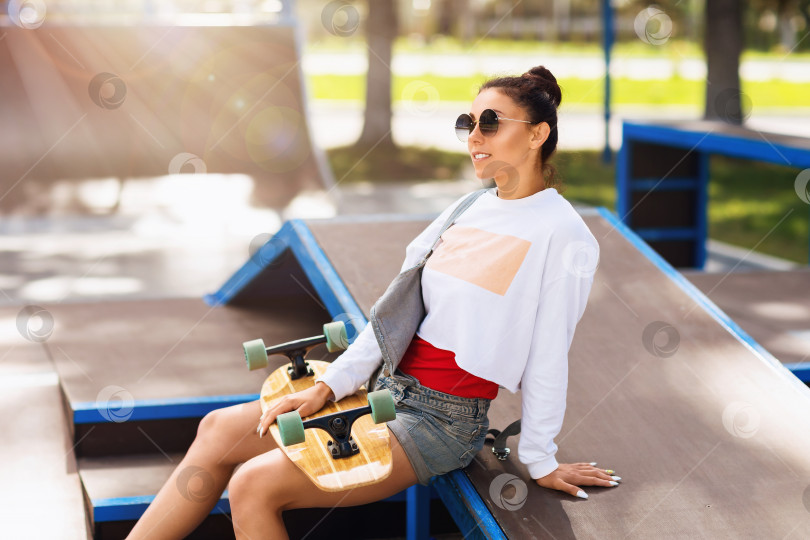 Скачать Молодая женщина с лонгбордом в руках отдыхает в парке для катания на скейтборде летним солнечным утром фотосток Ozero