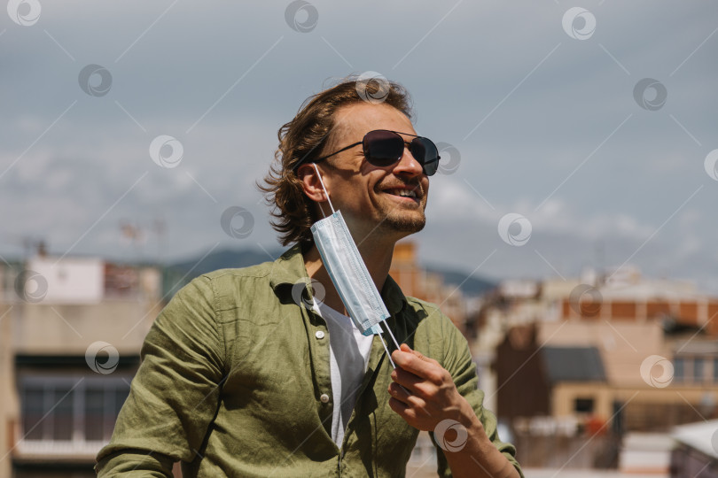 Скачать Мужчина в солнцезащитных очках радостно снимает медицинскую маску со своего лица в солнечный день на улице. Окончание карантина в связи с пандемией. фотосток Ozero