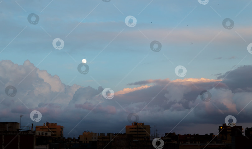 Скачать Пышные кучевые облака плывут по вечернему небу вместе с луной теплым летним вечером над городскими высотными зданиями. Концепция природного явления. Copyspace фотосток Ozero