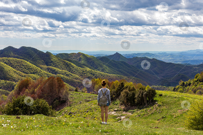 Скачать Девушка-путешественница с рюкзаком стоит на холме и любуется удивительным видом на горную долину, выбирая маршрут. Вид сзади. фотосток Ozero