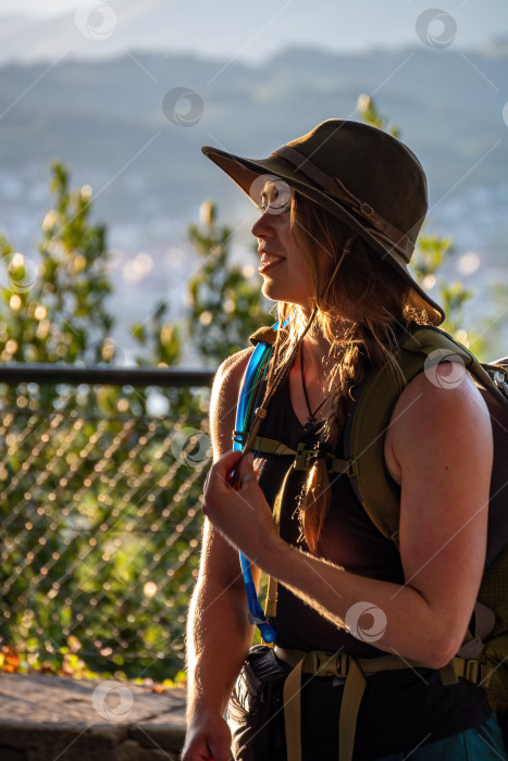 Скачать Портрет привлекательной улыбающейся американской девушки в оливковой шляпе с рюкзаком во время пешего похода в горы, наслаждающейся солнечным днем и хорошим настроением. фотосток Ozero