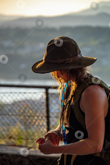 Скачать Американская девушка трогает кольцо на своем пальце. Стоит в оливковой шляпе с рюкзаком во время пешего похода в горы. фотосток Ozero