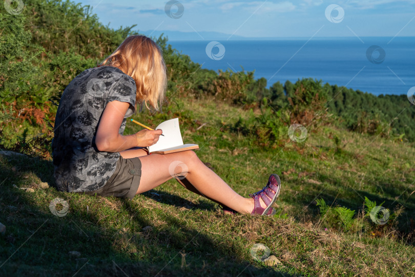Скачать Молодая светловолосая девушка записывает свои мысли в личный дневник во время пешего похода по горе с видом на море. Наслаждайтесь своим временем и моментами. Путешествия и отдых фотосток Ozero