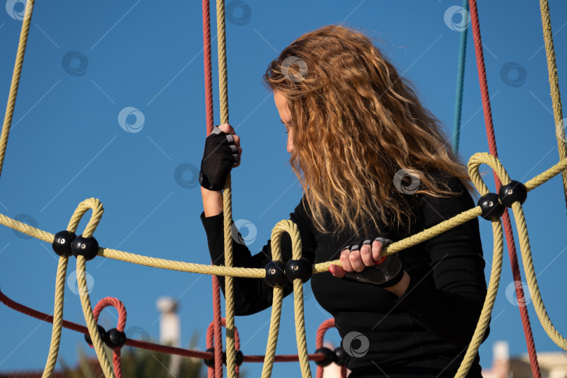 Скачать Активная кудрявая девочка в черной одежде играет с сеткой для скалолазания на детской площадке в парке. Молодая спортивная женщина развлекается в развлекательном центре фотосток Ozero