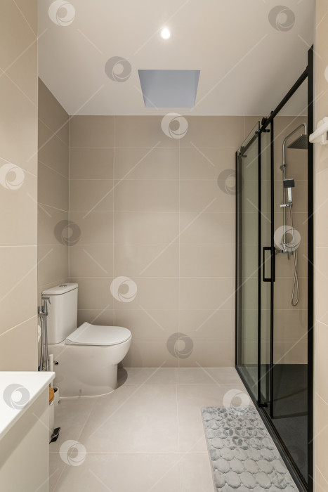 Скачать Вертикальный снимок просторной ванной комнаты с унитазом, застекленной душевой кабиной и умывальником с тумбой в бежевых тонах в квартире. Концепция практичного и лаконичного ремонта в ванной комнате фотосток Ozero