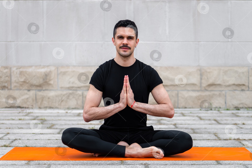Скачать Латиноамериканский гей практикует йогу на открытом воздухе в городе, сидя в позе йоги на оранжевом коврике, на фоне серой стены. фотосток Ozero