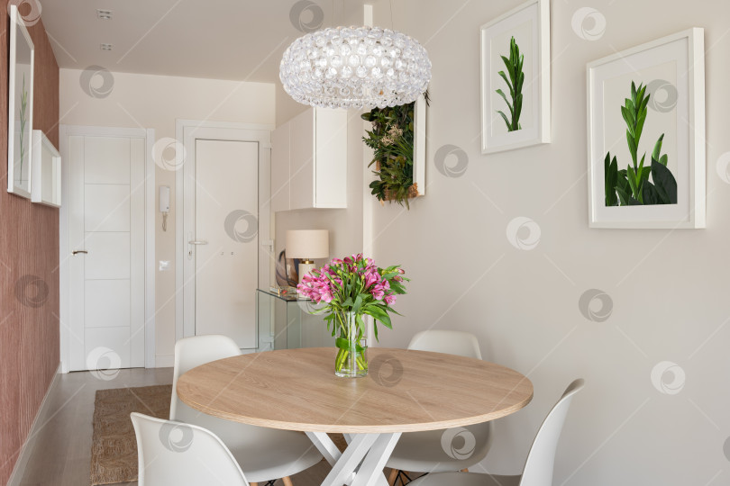 Скачать Прихожая-столовая с круглым деревянным столом, белыми стульями и картинами в интерьере современной квартиры. фотосток Ozero