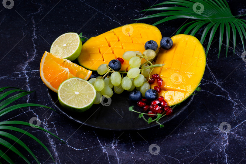 Скачать Ассорти из тропических фруктов подается на темном блюде: манго, виноград, лайм, апельсин, черника и гранатовые косточки на черном столе. фотосток Ozero