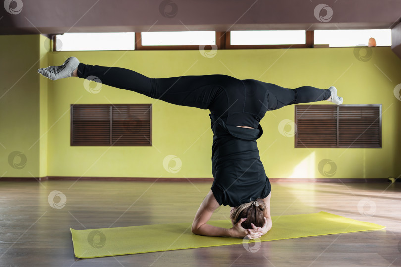 Скачать Молодая женщина, выполняющая упражнения ширшасана с самаконасаной, стойка на голове с опорой на локти, тренируется в черных леггинсах и футболке в студии на коврике фотосток Ozero