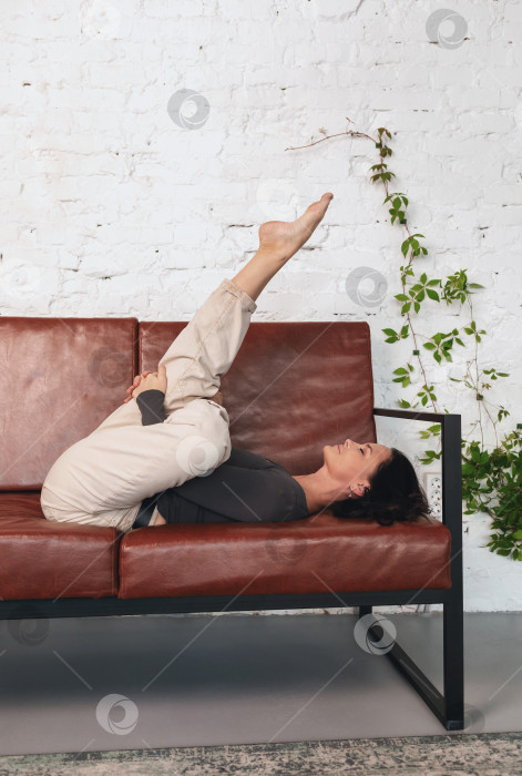 Скачать Привлекательная брюнетка выполняет вариацию упражнения Супта Экапада Агни Стамбхасана, практикуя йогу, лежа на диване в повседневной одежде фотосток Ozero