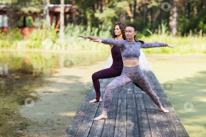 Скачать Две женщины, практикующие йогу утром на деревянном мосту в парке, выполняют упражнение вирабхадрасана, поза воина фотосток Ozero