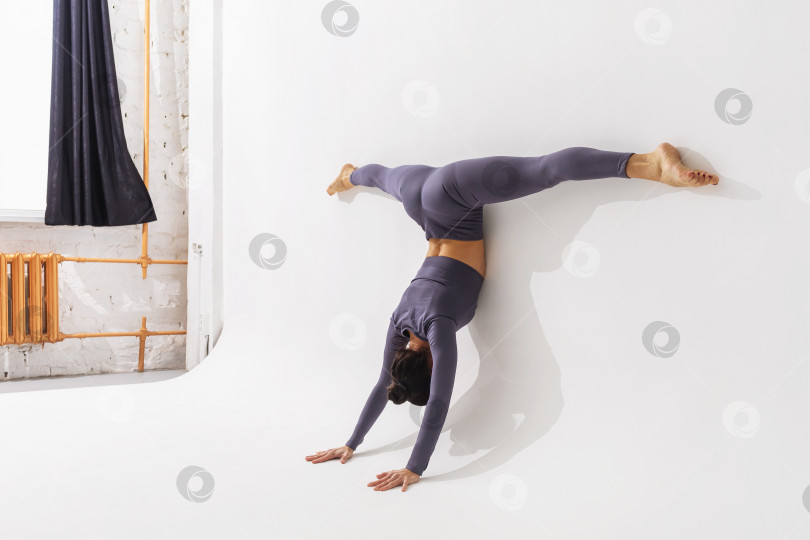 Скачать Женщина, практикующая йогу, выполняет упражнение Адхо Мукха врикшасана с Самаконасаной, стоит на руках с поперечным шпагатом, прислонившись телом к стене фотосток Ozero