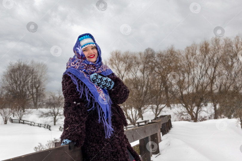 Скачать Добродушно смеющаяся русская женщина в народном костюме - шубе, шали, кокошнике и варежках у забора на фоне зимнего пейзажа фотосток Ozero