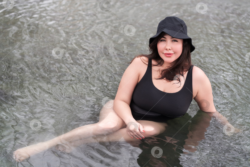 Скачать Крупногабаритная модель в черном купальнике и панаме расслабляется в открытом бассейне спа-курорта фотосток Ozero