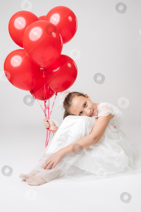 Скачать Романтичная девушка держит в руках много красных воздушных шаров, положила голову на колени, сидит на белом фоне фотосток Ozero