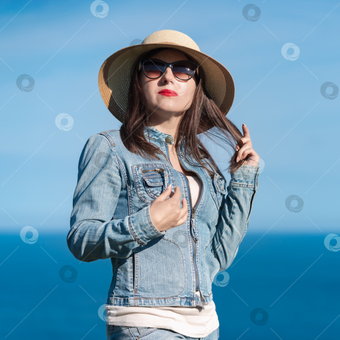 Скачать Портрет женщины в джинсовой куртке, джинсах, соломенной шляпе и солнцезащитных очках на фоне голубого неба, океана фотосток Ozero