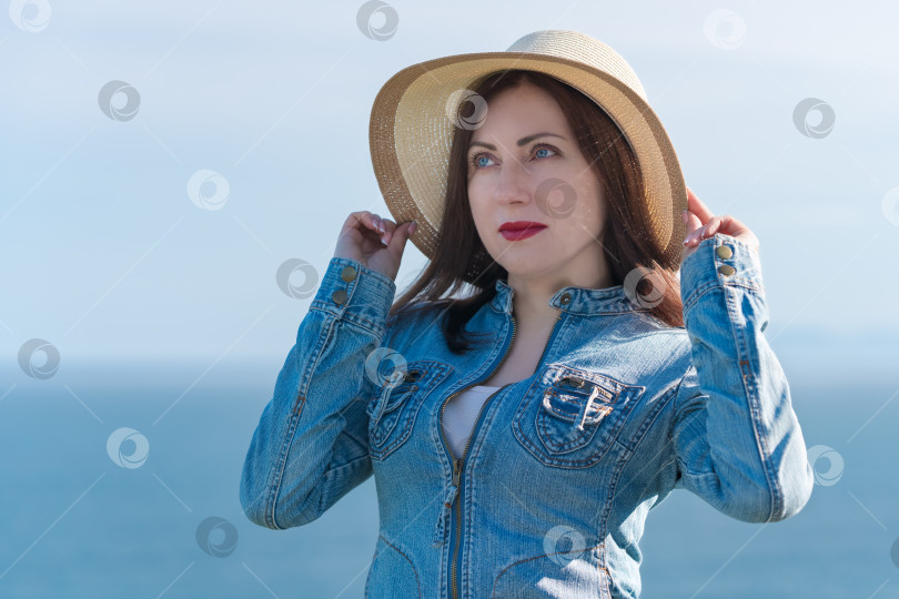 Скачать 40-летняя женщина-хипстер подняла руки и держит соломенную шляпу на голове, позируя на фоне неба и океана фотосток Ozero