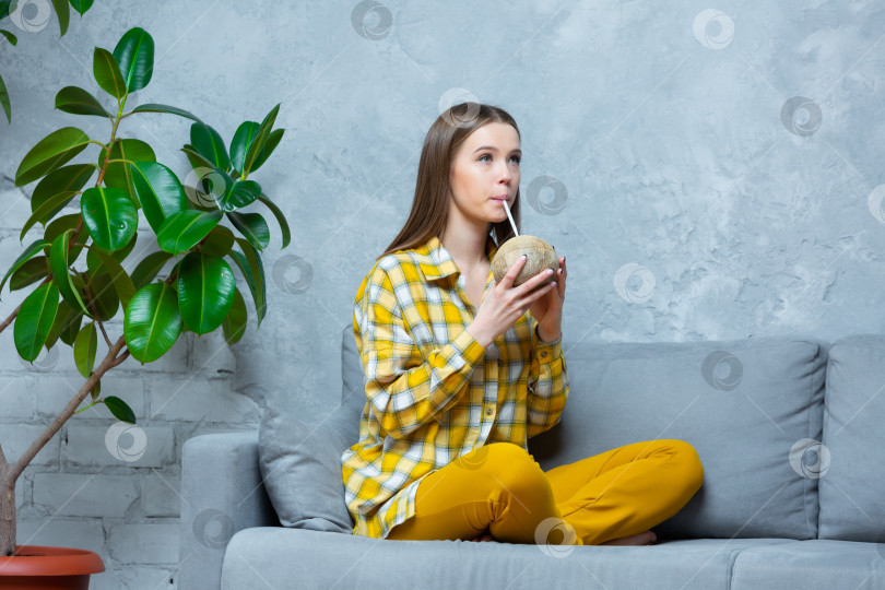 Скачать Фотография красивой молодой женщины, сидящей с ногами на диване и пьющей кокосовое молоко из кокоса через соломинку. Концепция релаксации, медитации, веганства, правильного питания фотосток Ozero