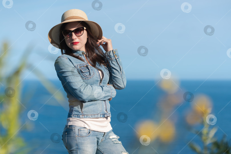 Скачать Женщина в солнцезащитных очках, соломенной шляпе, джинсовой куртке и джинсах позирует на фоне голубого неба и океана фотосток Ozero