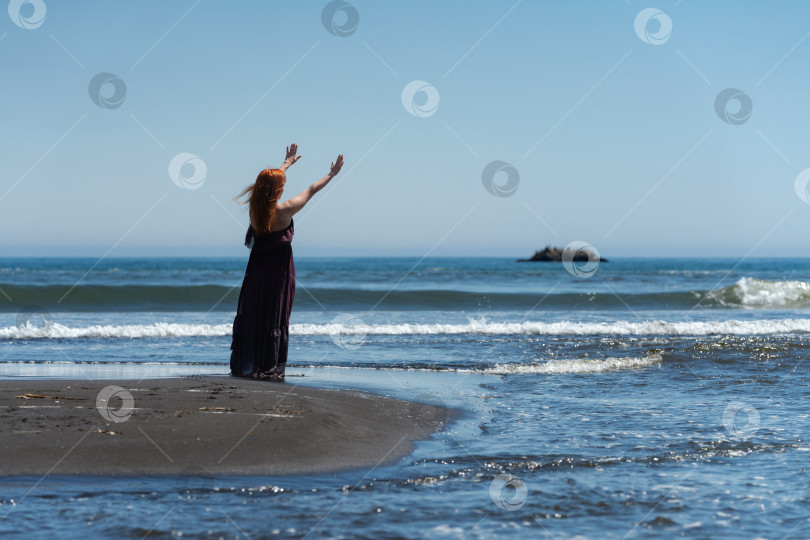 Скачать Вид сзади женщина в длинном платье, стоящая на песчаном пляже с поднятыми руками, простирающимися к небу и морю фотосток Ozero