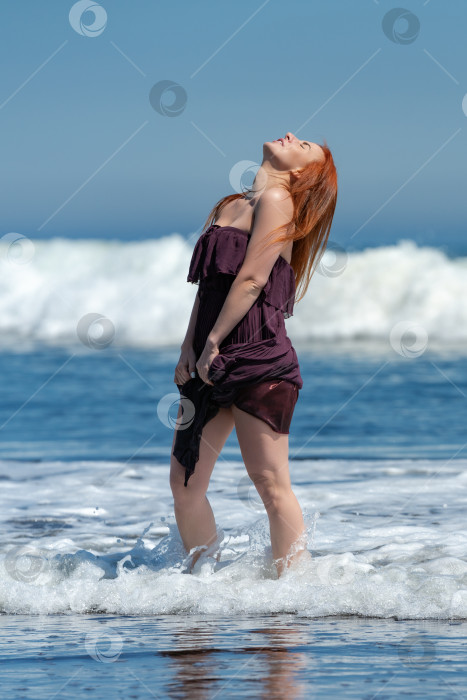 Скачать Счастливая женщина в длинном платье с приподнятым подолом стоит по щиколотку в воде на пляже, подняв голову вверх фотосток Ozero