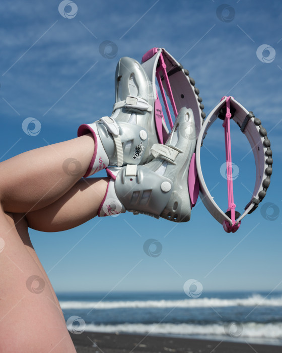 Скачать Женщина со скрещенными ногами в воздухе, обутая в розовые ботинки Kangoo Jumps, во время занятий фитнесом на открытом воздухе на фоне голубого неба и пляжа на морском побережье фотосток Ozero