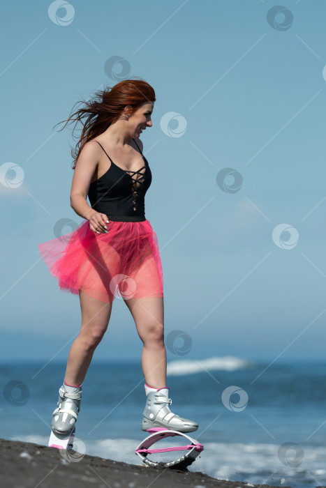 Скачать Спортивная женщина в ботинках Kangoo Jumps, цельном купальнике и короткой розовой юбке бегает и прыгает по черному песчаному пляжу во время тренировки по фитнесу фотосток Ozero