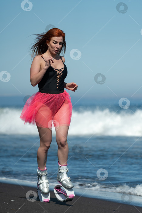 Скачать Спортсменка в ботинках Kangoo Jumps, цельном купальнике и короткой юбке бегает и прыгает по черному песчаному пляжу во время занятий аэробикой и фитнесом фотосток Ozero