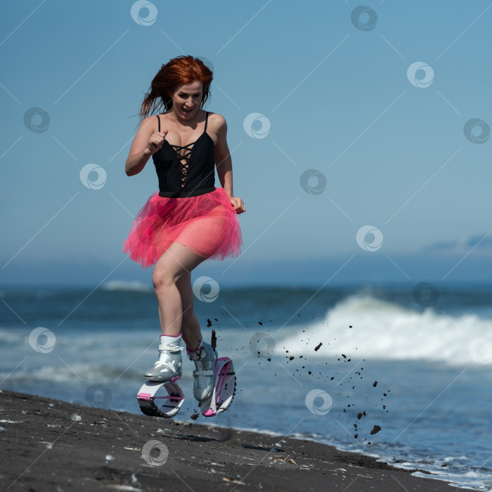 Скачать Женщина в спортивных ботинках Kangoo Jumps, черном цельном купальнике и короткой розовой юбке прыгает и бегает по песчаному пляжу во время занятий фитнесом на открытом воздухе фотосток Ozero