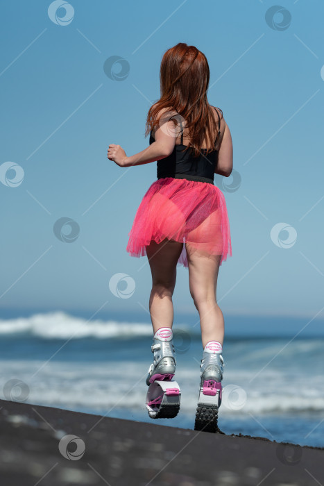 Скачать Вид сзади на спортивную женщину в спортивных ботинках Kangoo Jumps, короткой розовой юбке, купальнике, бегущую и прыгающую по пляжу во время занятий фитнесом на открытом воздухе фотосток Ozero