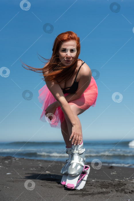 Скачать Рыжеволосая подтянутая женщина в ботинках Kangoo Jumps, купальнике и короткой юбке позирует на песчаном пляже во время тренировки по фитнесу на открытом воздухе летним днем фотосток Ozero