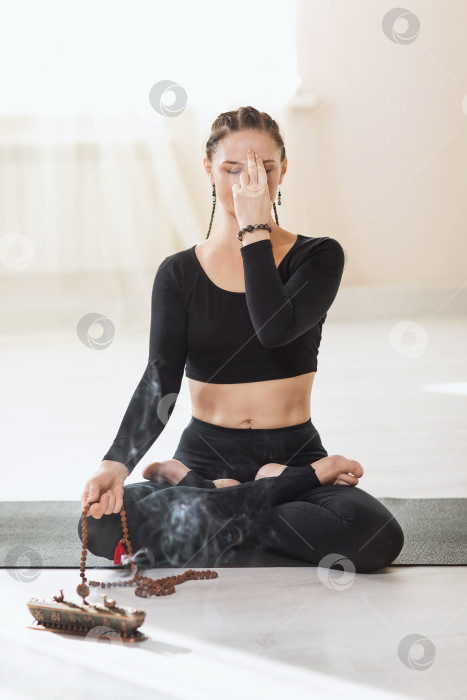 Скачать Женщина в черной спортивной одежде, практикующая йогу, сидя на гимнастическом коврике в позе лотоса, занимается медитацией и дыхательными упражнениями пранаяма. Инструктор показывает символ мудры рядом с лицом фотосток Ozero