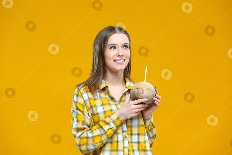 Скачать Снимок привлекательной молодой улыбающейся женщины, смотрящей в сторону и держащей в обеих руках кокосовый орех с соломинкой, выделенной на желтом фоне. Концепция здорового образа жизни, правильное питание, диета, веган, вегетарианец фотосток Ozero