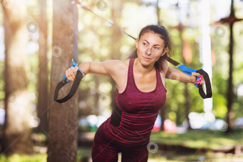 Скачать Молодая женщина делает упражнения в парке с помощью фитнес-ремней, прикрепленных к дереву. Спортсмен тренирует мышцы груди теплым летним утром фотосток Ozero