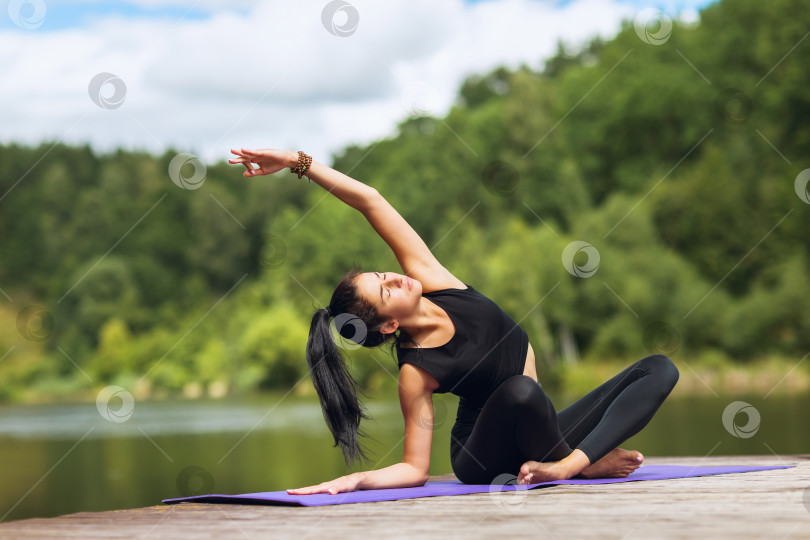 Скачать Молодая женщина в черной спортивной одежде на деревянном мосту у озера практикует асаны йоги фотосток Ozero