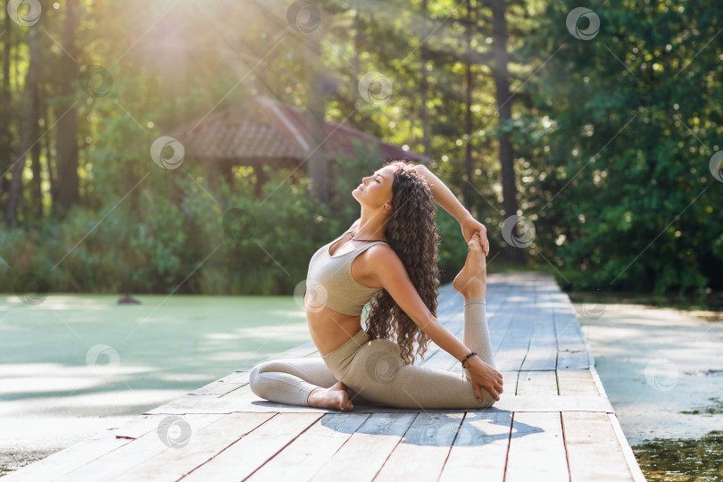 Скачать Женщина в леггинсах и коротком топе, практикующая йогу, сидящая на коврике в парке, выполняющая упражнение эка пада раджакапотасана, поза голубя фотосток Ozero