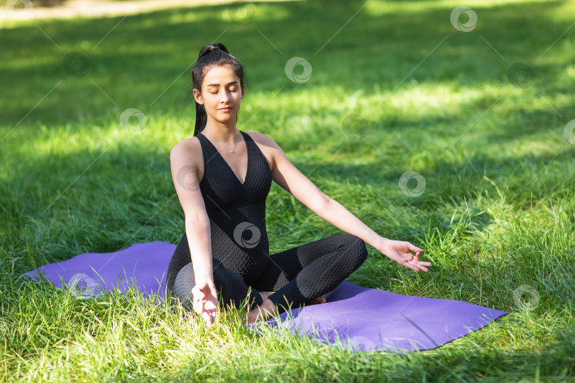 Скачать Молодая беременная женщина медитирует, сидя в позе лотоса с джняна мудрой, занимается йогой на коврике в парке в черном спортивном цельном комбинезоне фотосток Ozero