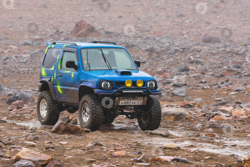 Скачать Японский внедорожник Suzuki Jimny едет по дороге скалистых гор на фоне вулканического пейзажа. Активный отдых, туристические направления, поездка по бездорожью в дождливую погоду фотосток Ozero