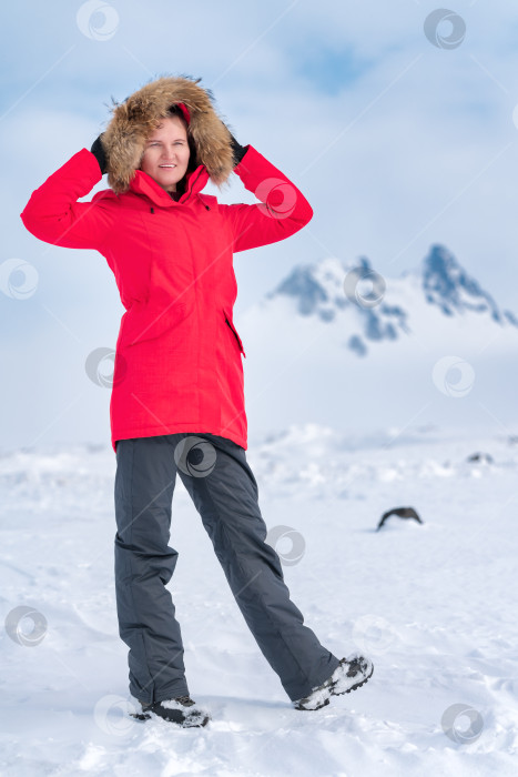 Скачать Женщина-альпинист, одетая в красную зимнюю куртку, держит капюшон над головой, стоит в зимних горах фотосток Ozero